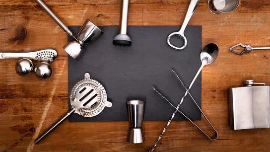 Stainless steel bar tools: jigger, strainer, bar spoon, muddler and ice tongs, laid out around a slate board on a warm wooden surface.