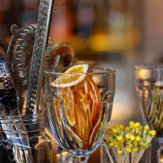 Dried orange and lemon wheels standing in a cut-glass tumbler beside bar tools, lit warm against a softly blurred back bar.